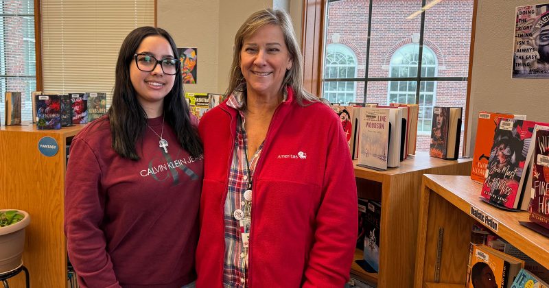 A caucasian woman in a red jacket, Wendy, stands by Isa, a latino teen girl, in a library. Both are smiling