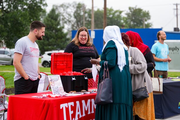 A man and a woman stand outside at a table with a TeamMates table cloth speaking to a woman wearing a headscarf